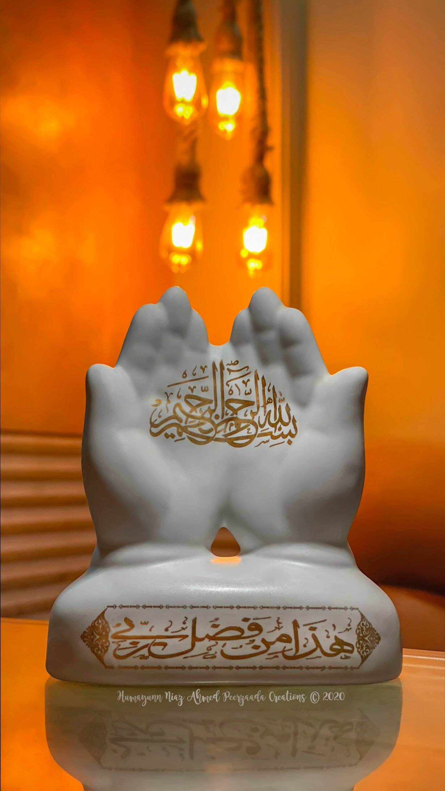 Hands sculpture with Arabic calligraphy “Bismillah ir-Rahman ir-Rahim” placed before glowing warm hanging lights — symbolizing prayer, gratitude, and divine blessings.