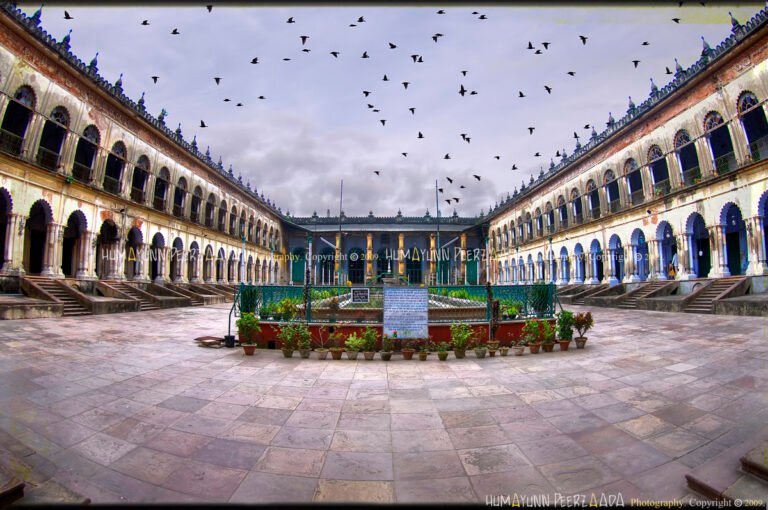 Wide-angle photograph of the grand courtyard of Hoogly Imambara in West Bengal, India — showcasing symmetrical arched corridors, historic architecture, and a flock of birds soaring in the dramatic sky.