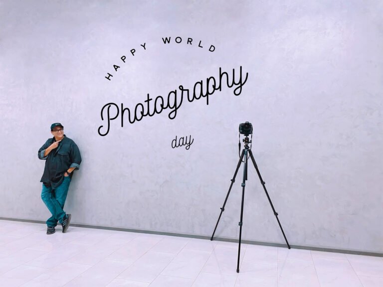 Humayunn Peerzaada standing beside a tripod in front of a wall with the words “Photography” written on it — celebrating World Photography Day.