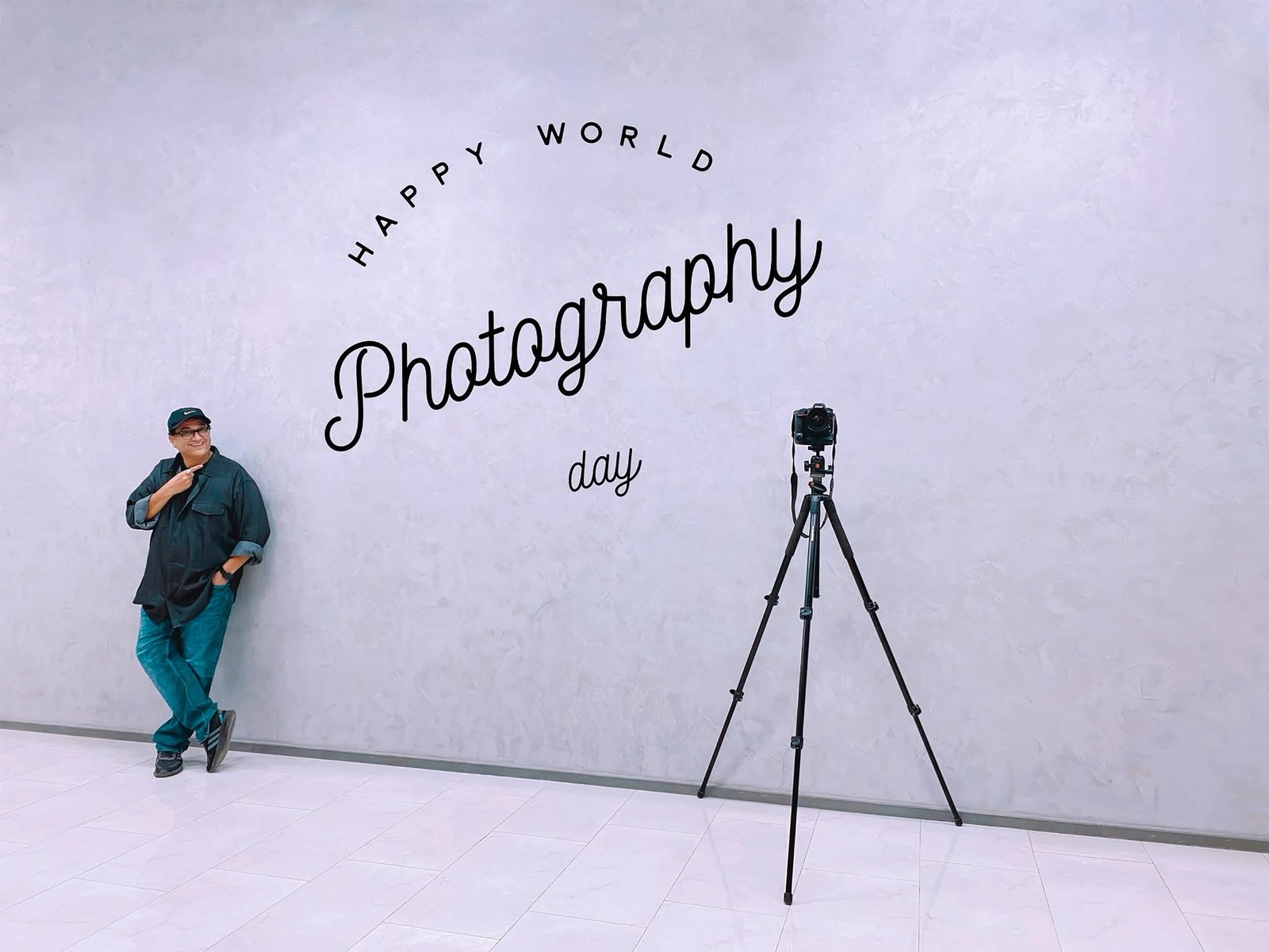Humayunn Peerzaada standing beside a tripod in front of a wall with the words “Photography” written on it — celebrating World Photography Day.