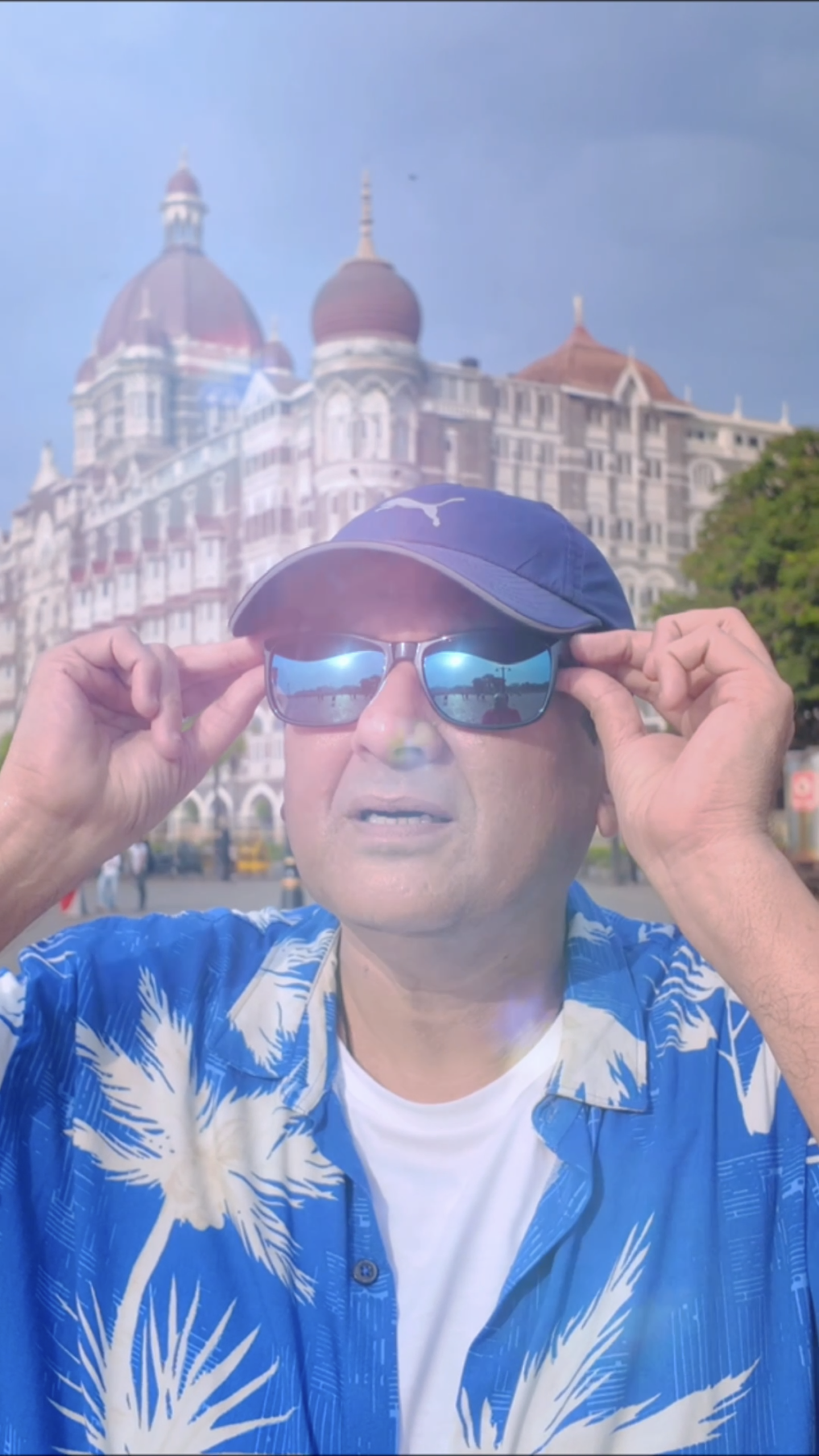 Man wearing a blue tropical shirt, cap and reflective sunglasses in front of the Taj Mahal Palace Hotel during a Gateway of India Mumbai travel vlog.